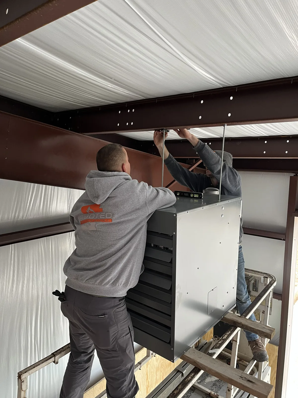 Two technicians hang a rectangular indoor HVAC unit from steel framing in a building under construction