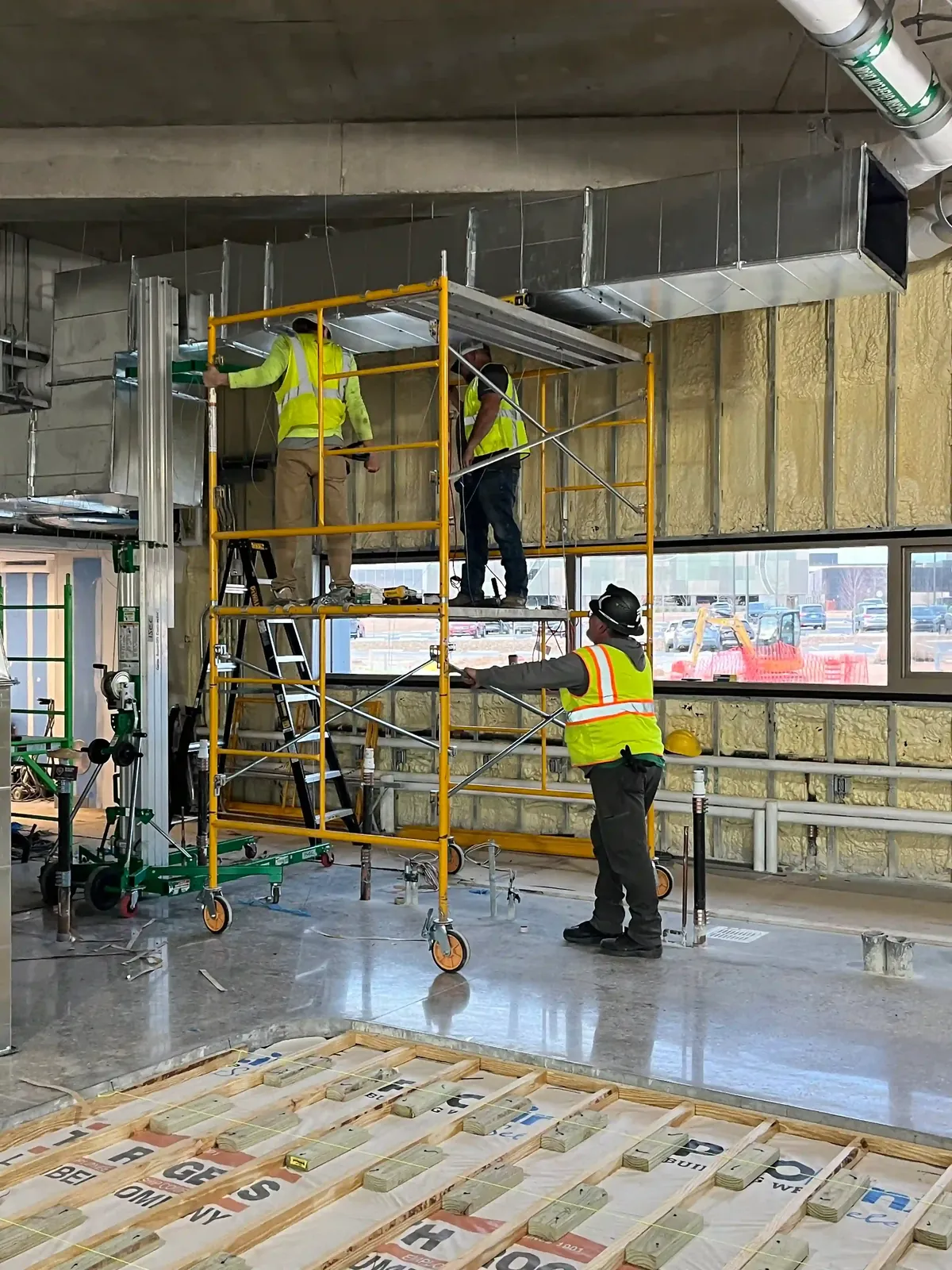 Technicians in high visibility vests on a rolling scaffold installing overhead ductwork