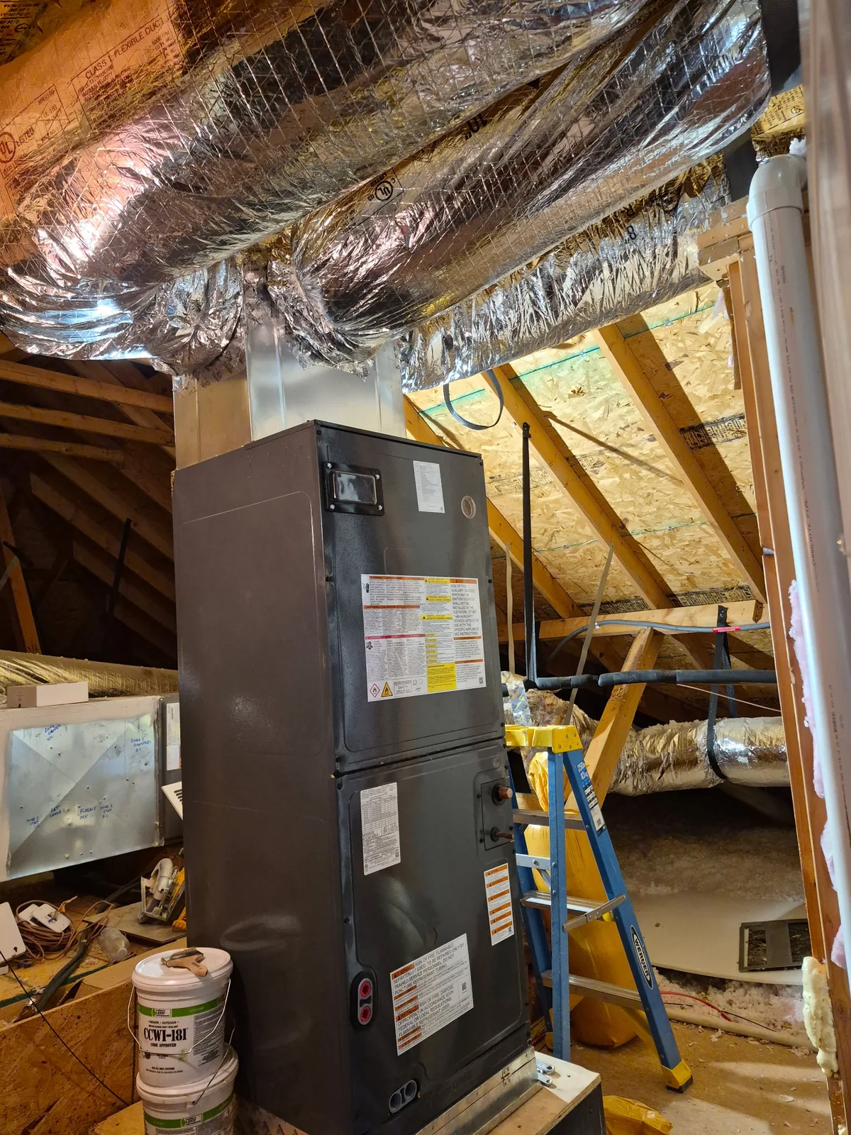 Upright air handler in an unfinished attic, under foil wrapped supply ducts, with a step ladder beside it
