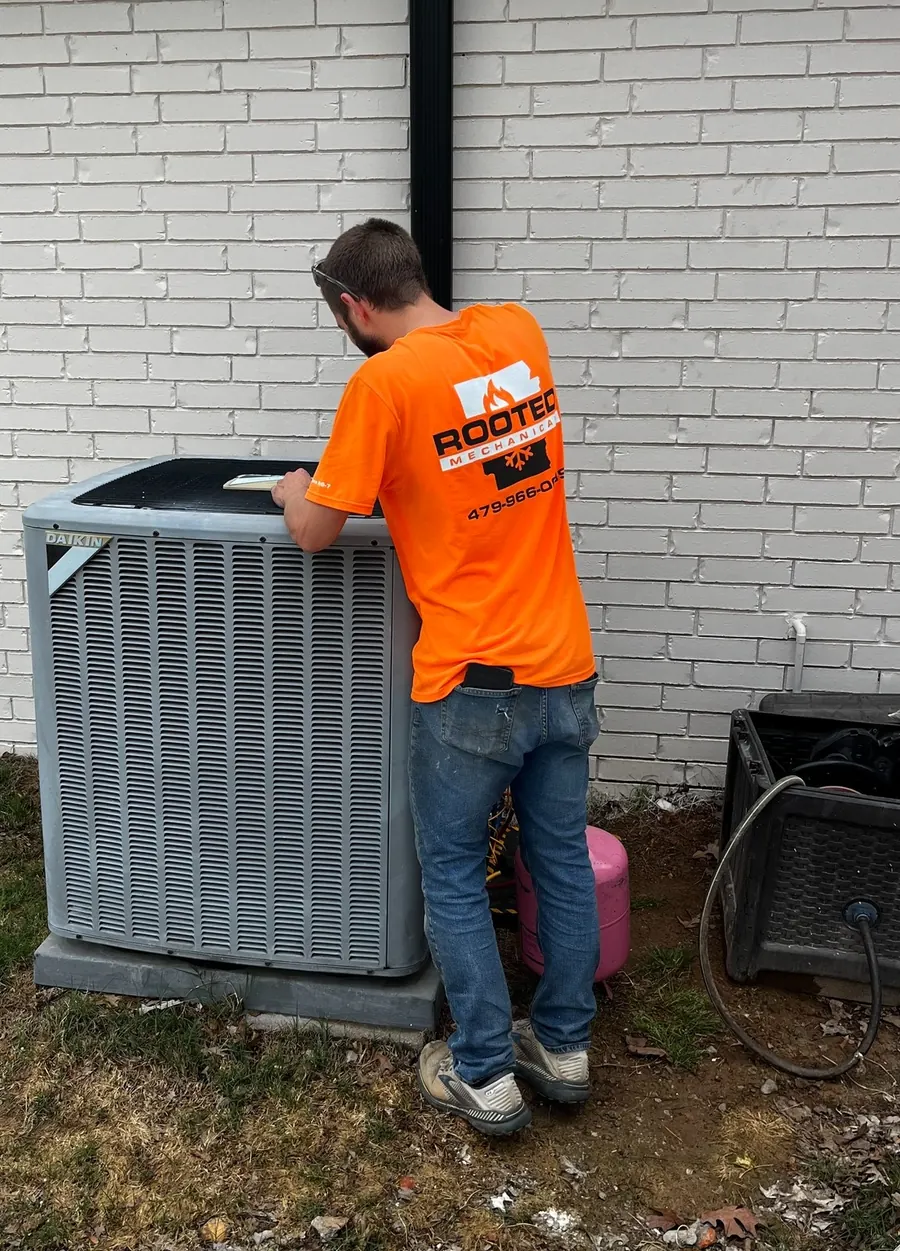 Rooted Mechanical Heating & Air technician in an orange company shirt working at an outdoor HVAC unit beside a white brick wall