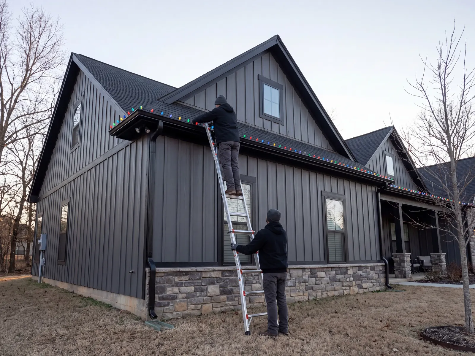 Two installers hanging multicolor C9 lights on a dark board-and-batten house, one on an extension ladder at the gutter and one steadying it