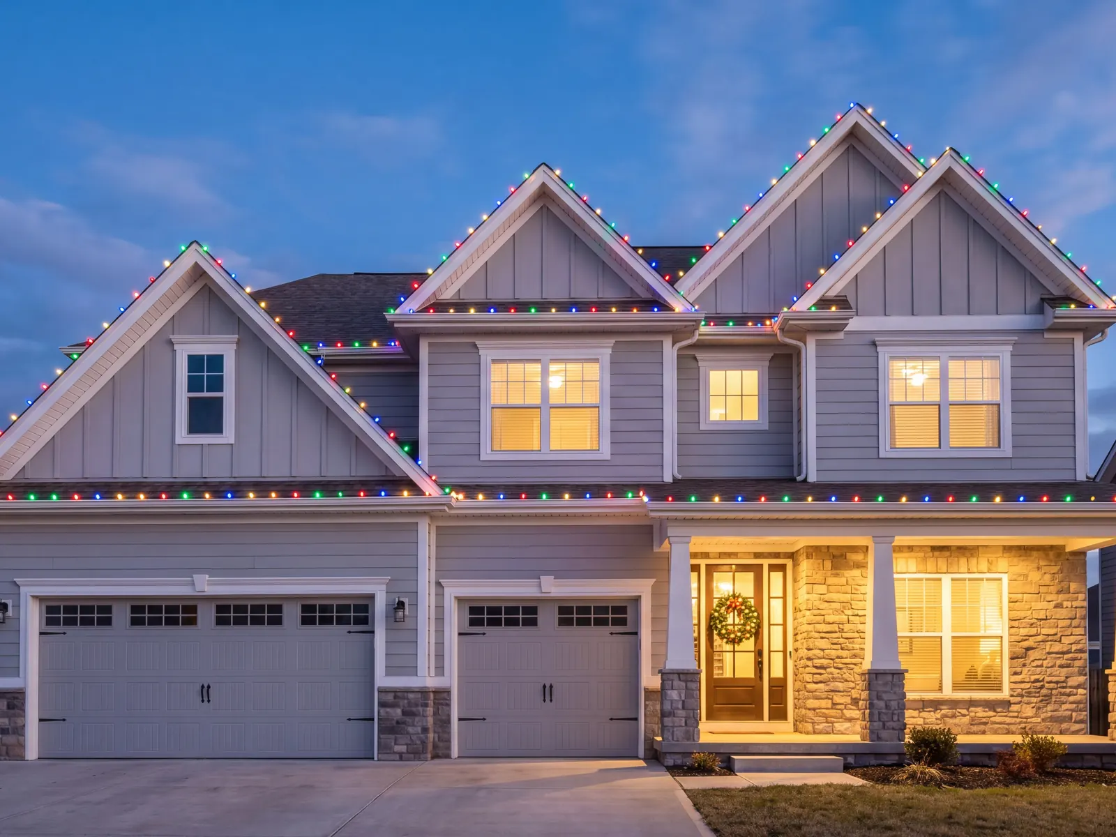 Two-story home at dusk with multicolor Christmas lights on three gables and a wreath on the front door