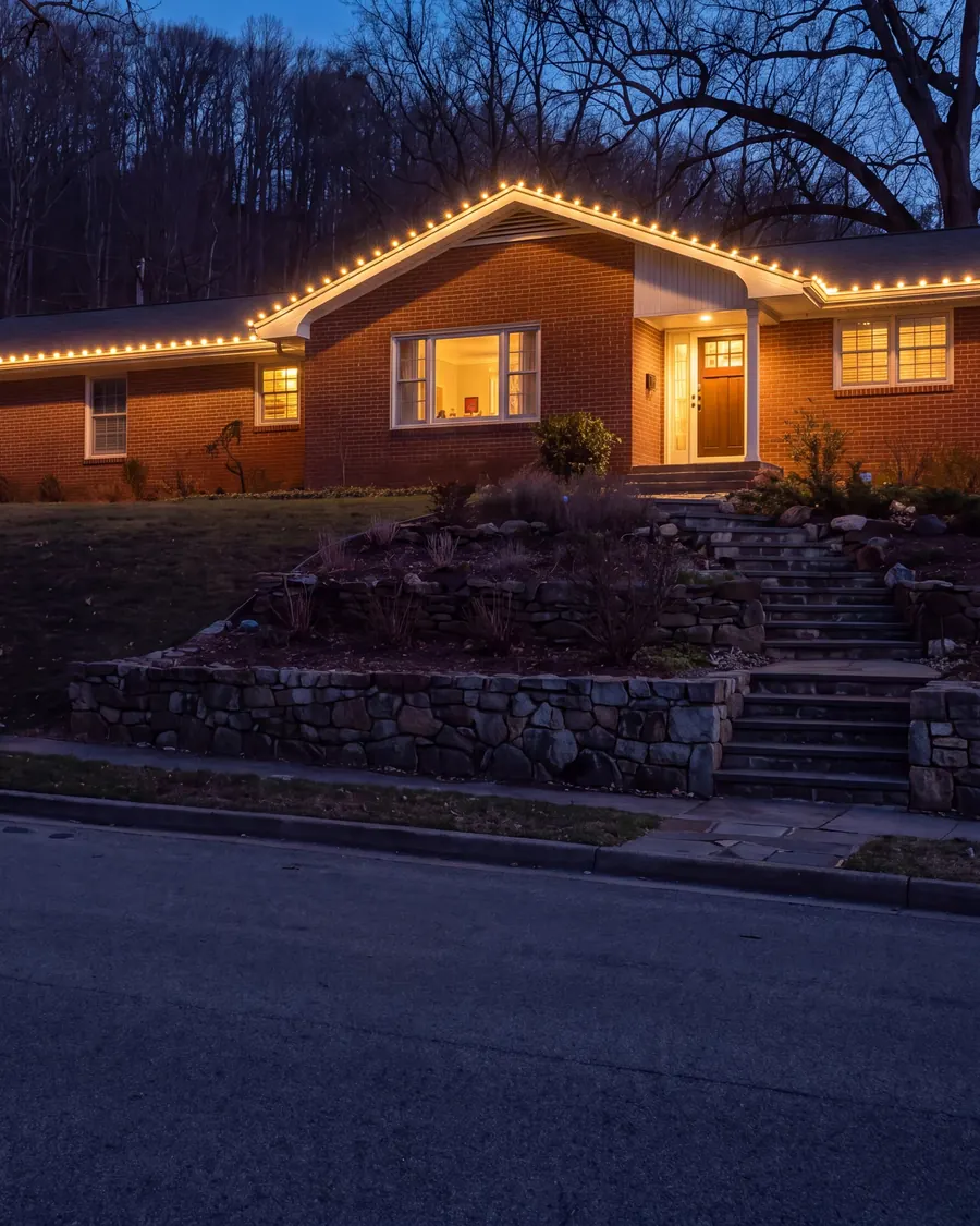 Brick ranch on a hillside at blue hour, warm-white lights along the roofline, two lit shrubs, stone steps and bare trees above