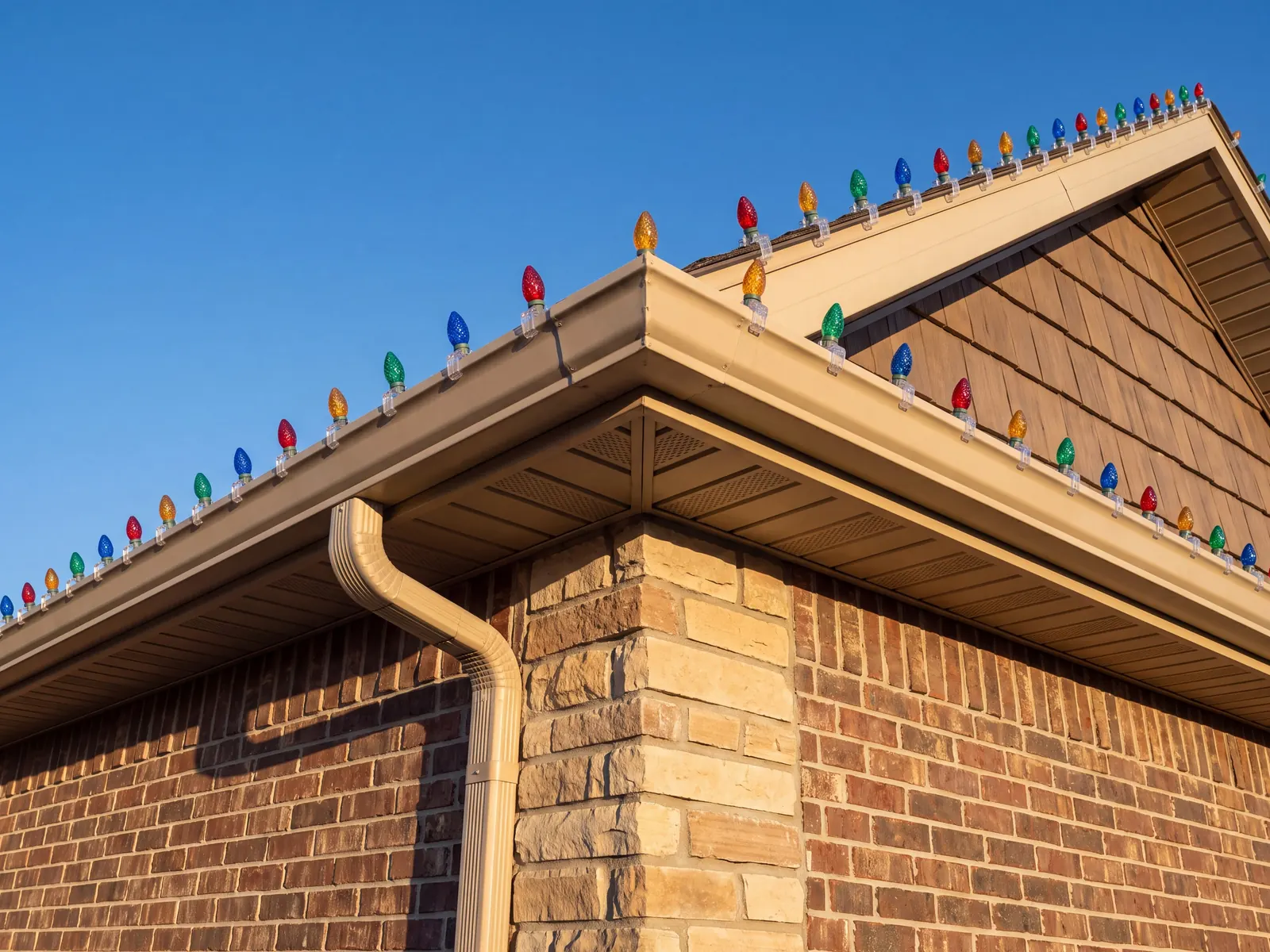 Multicolor C9 bulbs clipped along the gutter and gable of a brick and stone house against a clear blue sky