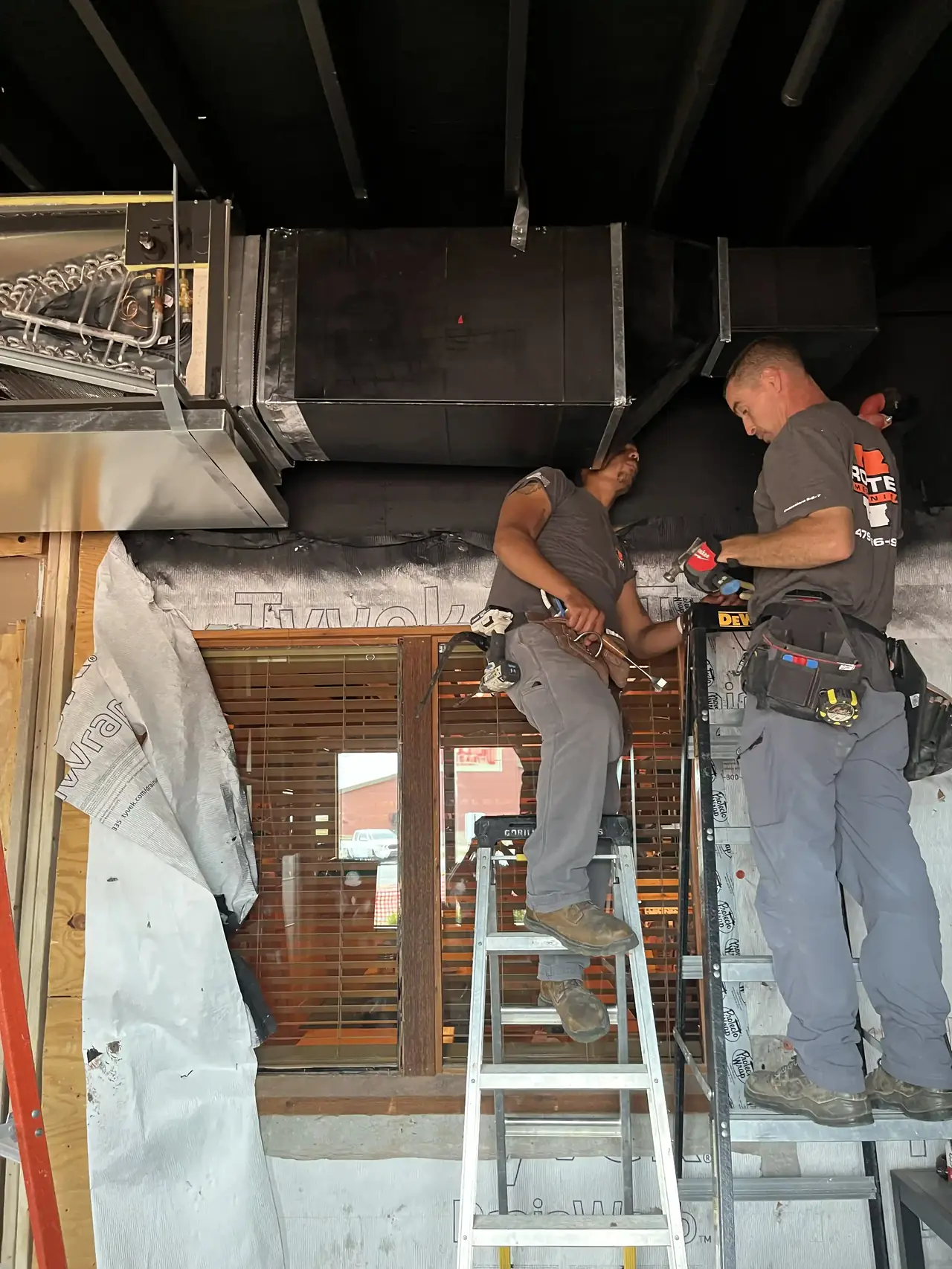 Two Rooted Mechanical Heating & Air technicians on ladders installing an overhead indoor HVAC unit and ductwork in a building under renovation