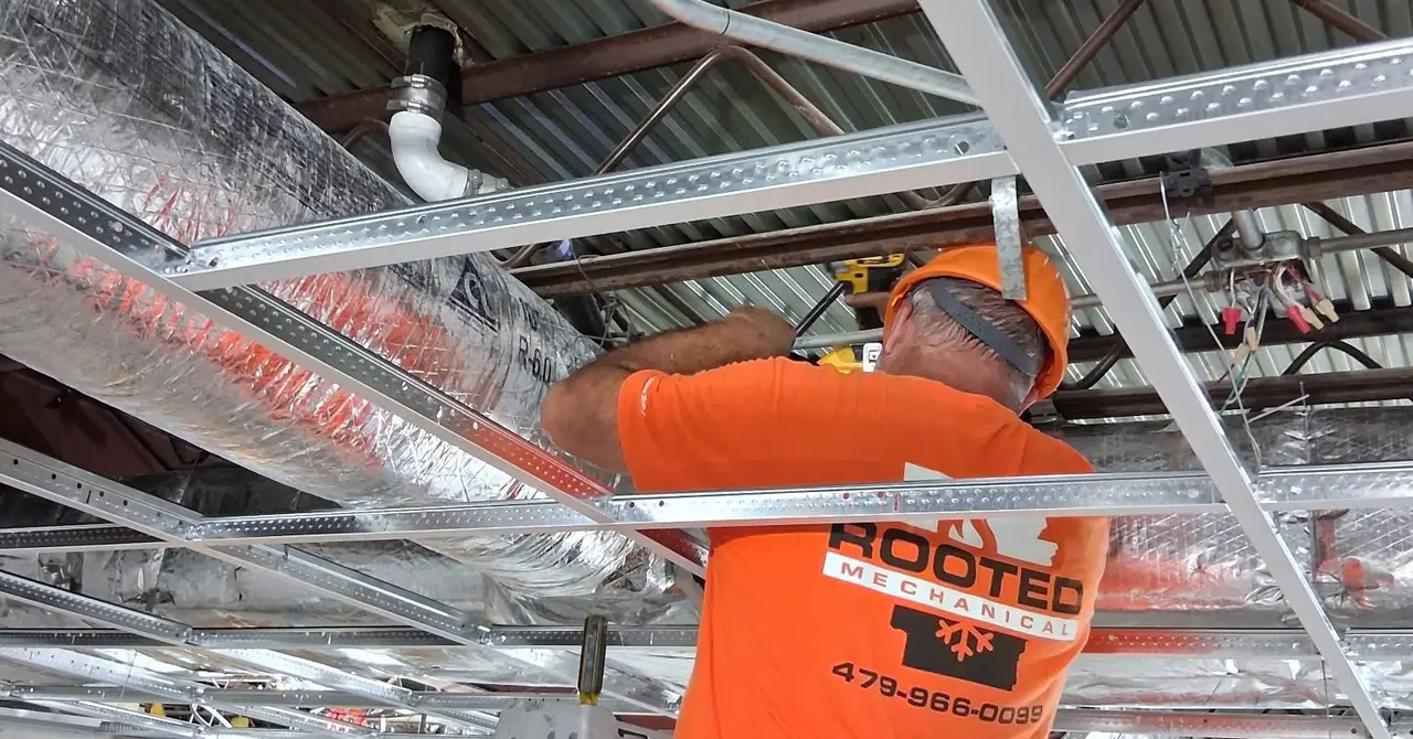 Rooted Mechanical Heating & Air technician in a hard hat installing insulated ductwork above a commercial drop-ceiling grid
