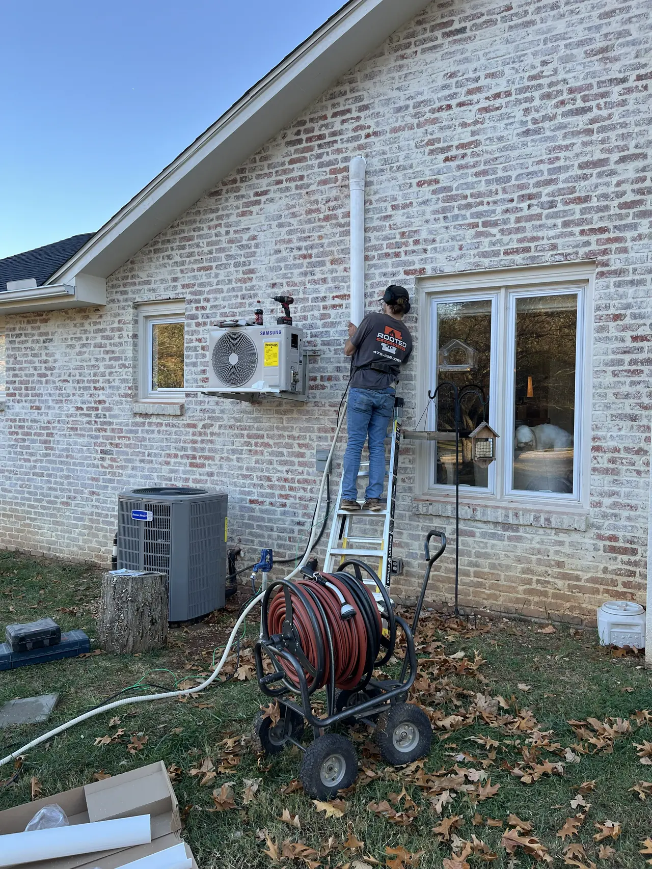 Technician on a ladder installing a ductless mini split indoor unit on the side of a painted brick home