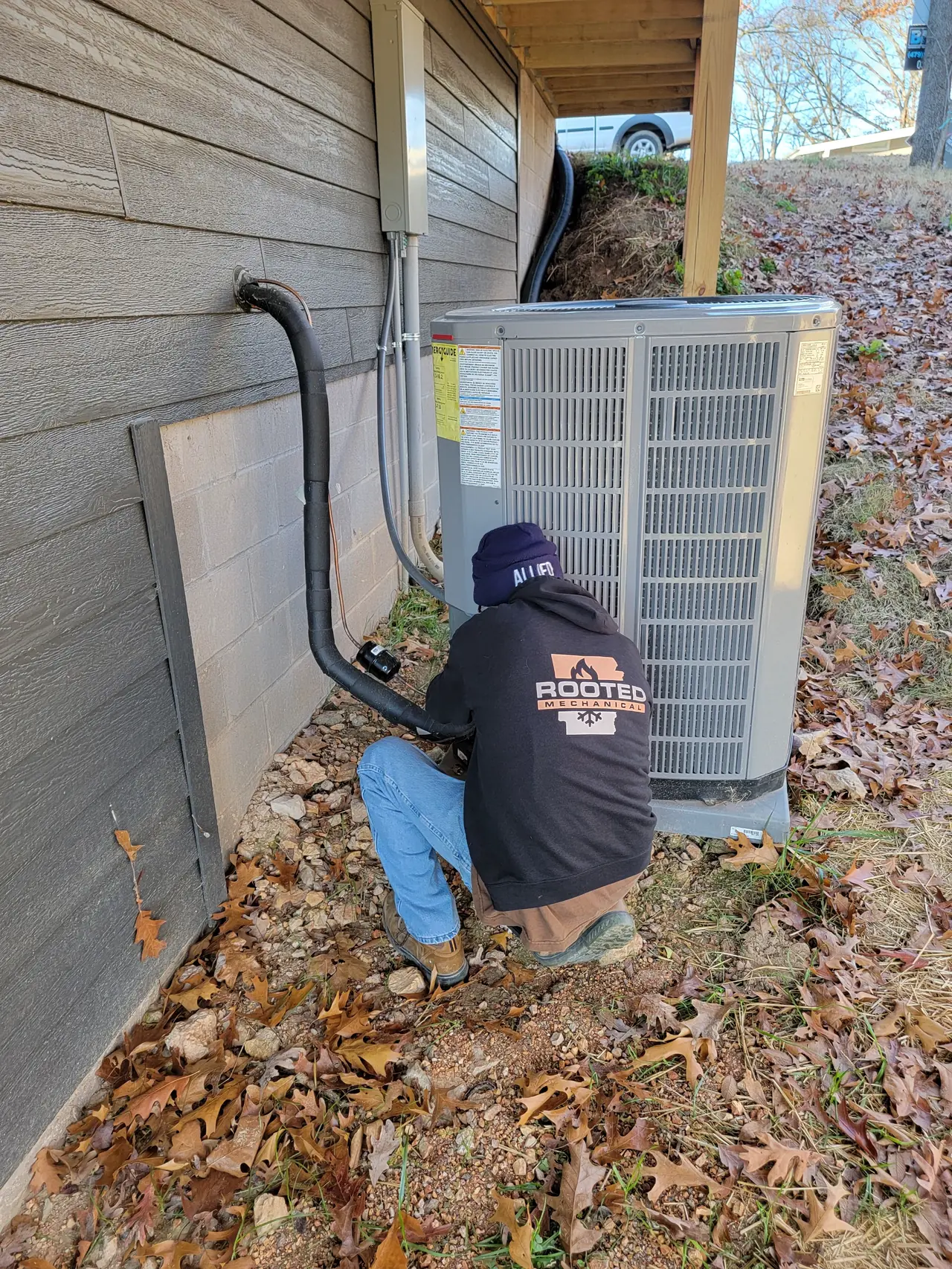 Rooted Mechanical Heating & Air technician in a black company hoodie crouched at an outdoor HVAC unit beside a gray-sided house