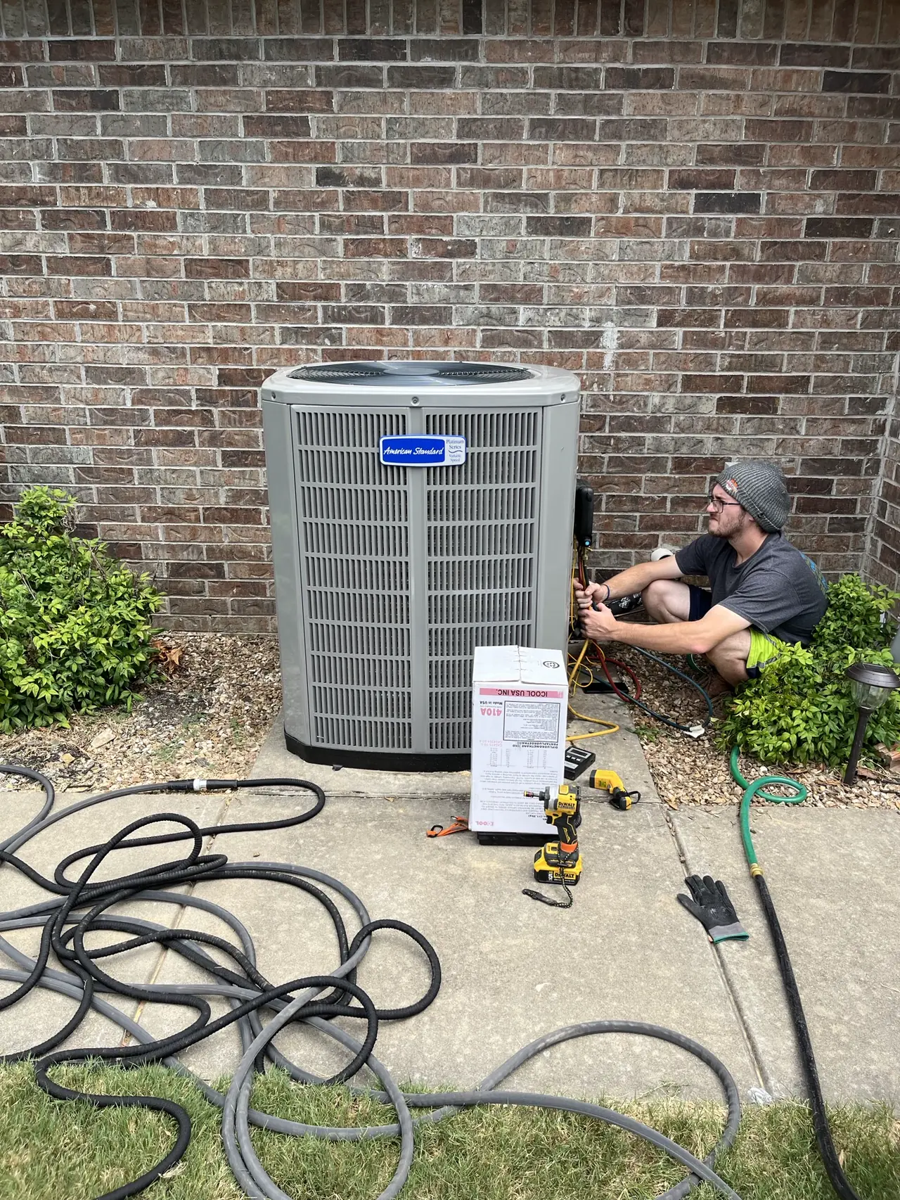 Technician working beside a new outdoor HVAC unit at a brick wall, with the unit's shipping box and tools nearby