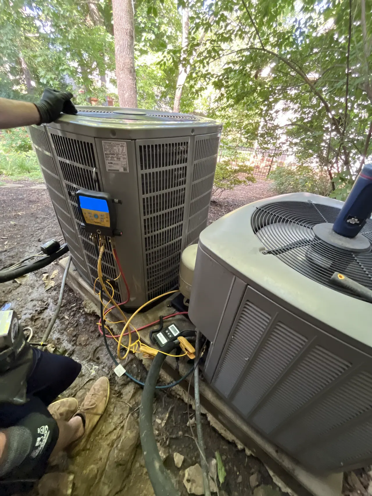 Close view of two outdoor HVAC units in a wooded yard, one with a digital gauge on its service panel