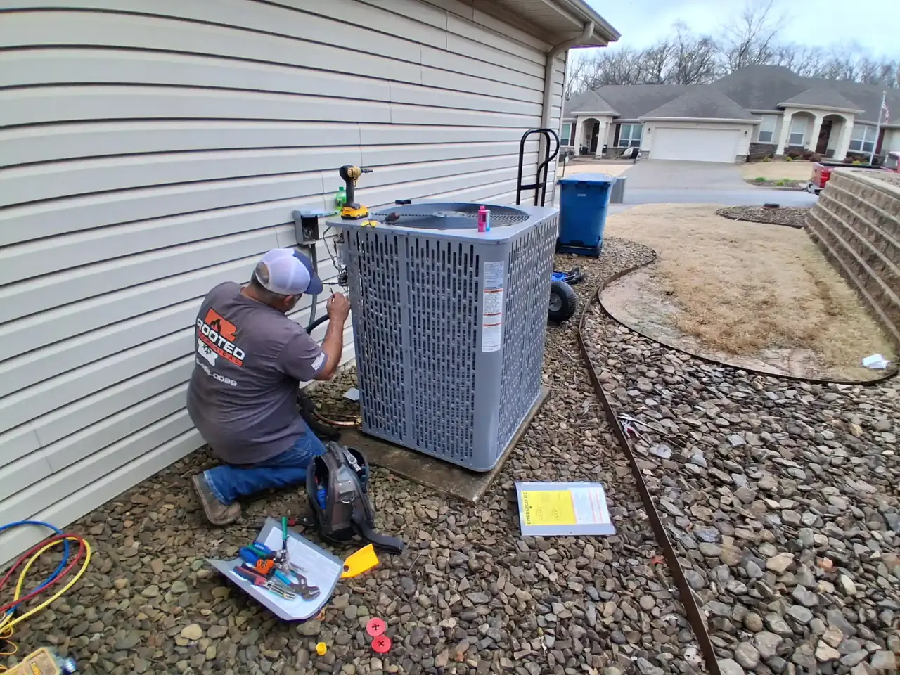 Rooted Mechanical Heating & Air technician kneeling at an outdoor HVAC unit beside a vinyl-sided home, with gauges and tools