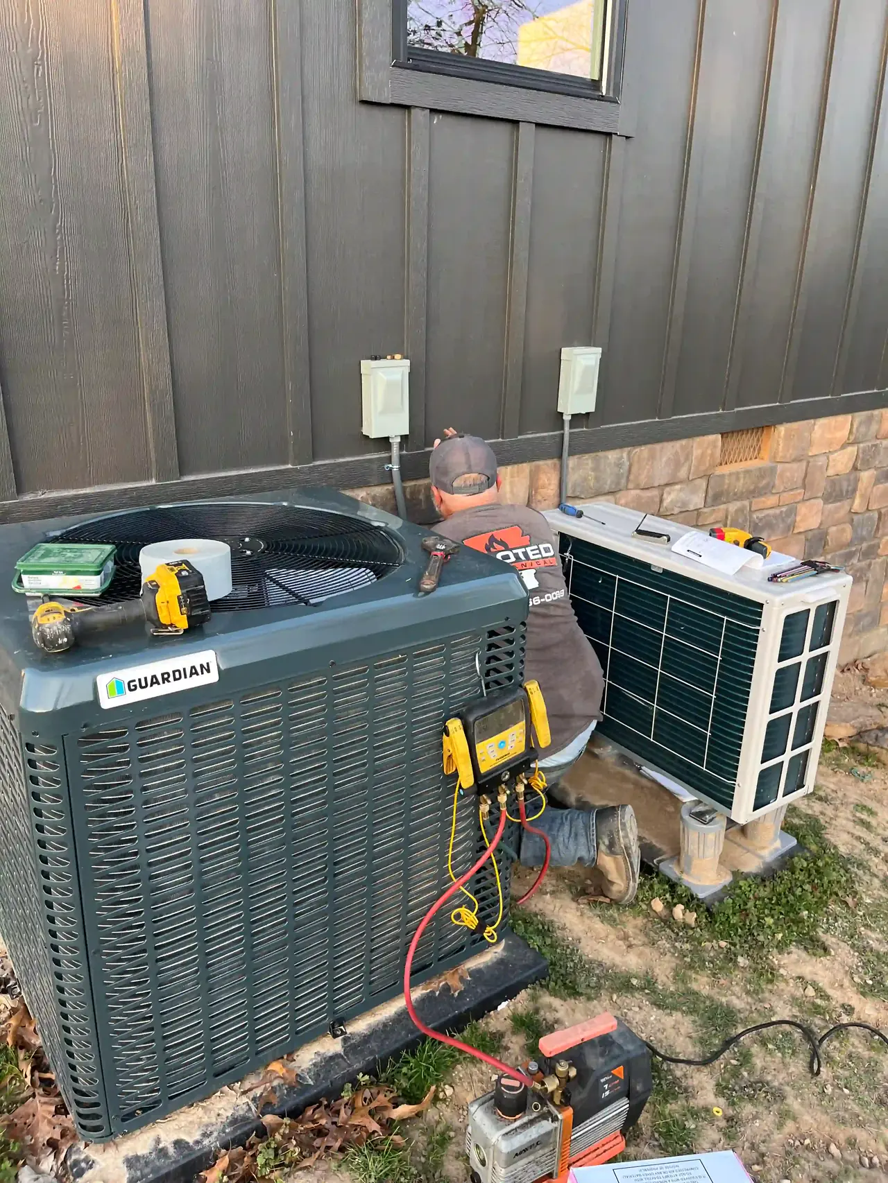 Rooted Mechanical Heating & Air technician kneeling between an outdoor HVAC unit fitted with refrigerant gauges and a ductless mini split outdoor unit