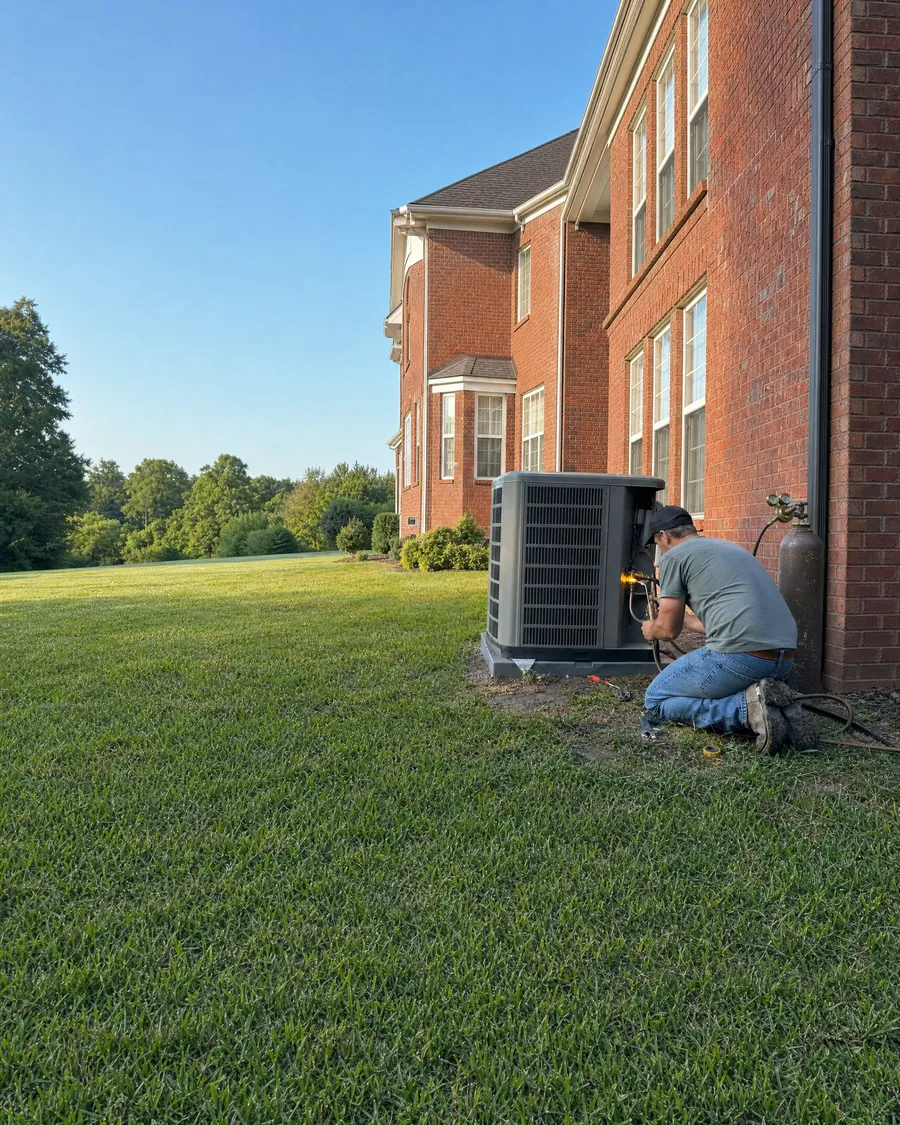 A technician brazes a copper line set on a new outdoor air conditioner beside a brick house.