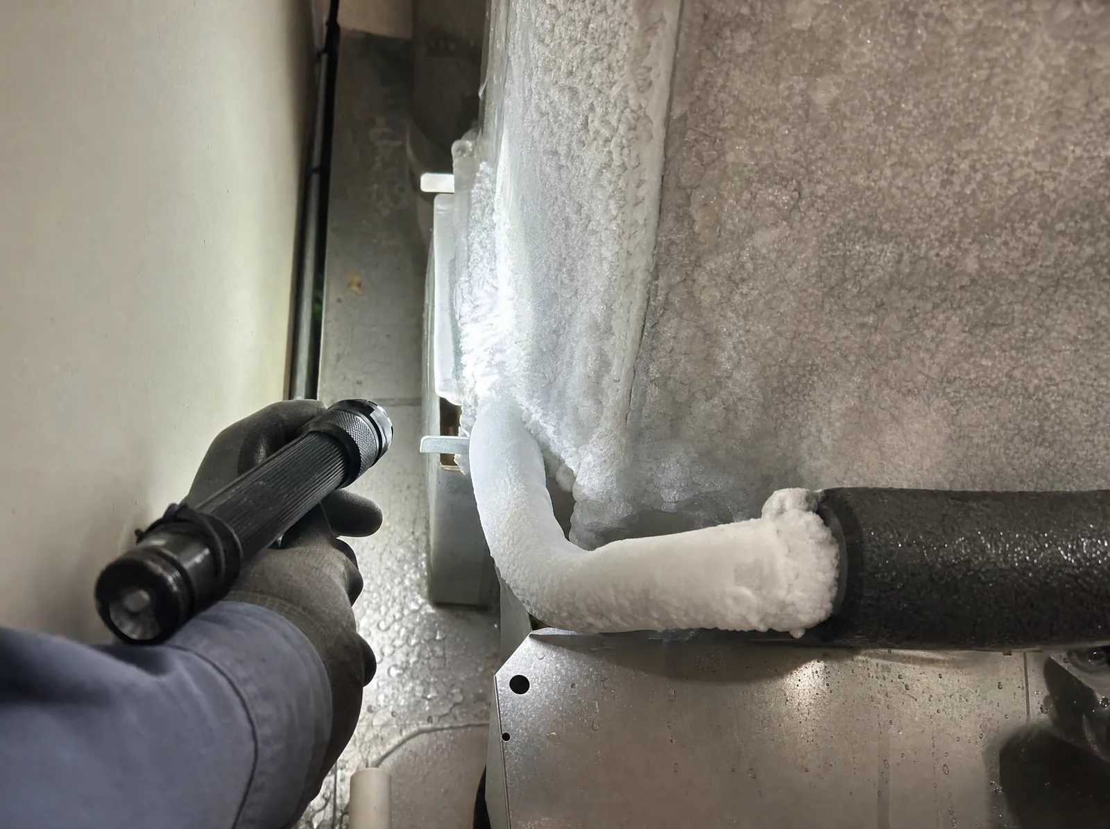 A gloved hand holds a flashlight on a refrigerant line covered in frost.
