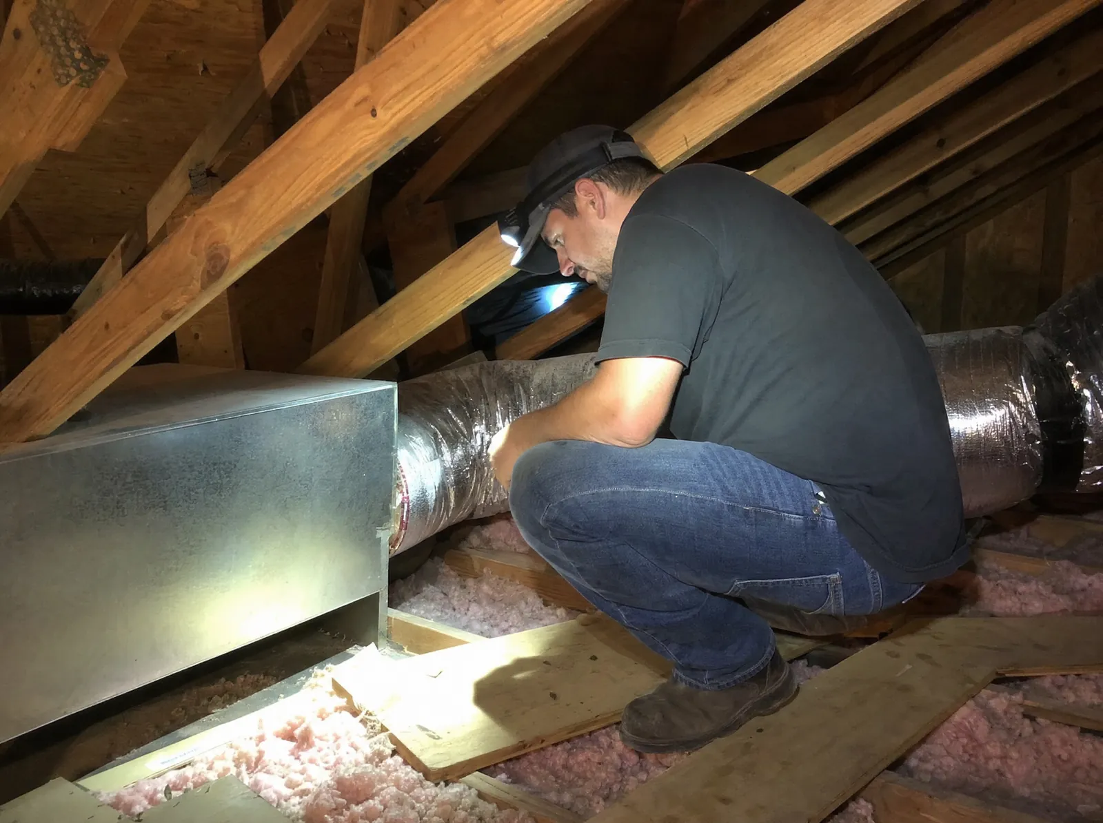 HVAC technician wearing a headlamp inspecting flexible duct and a sheet metal supply plenum in an insulated attic