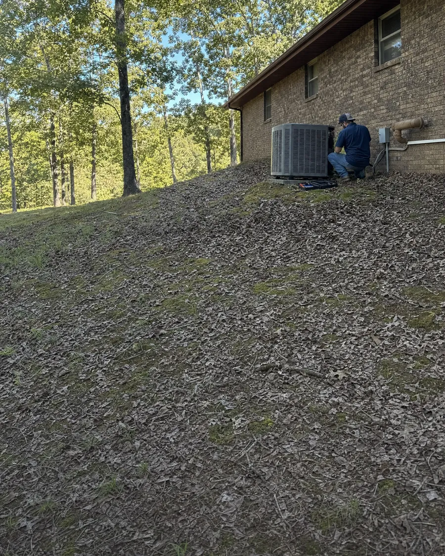 HVAC technician kneeling at an outdoor air conditioner beside a brick ranch home on a wooded hillside lot covered in fallen leaves