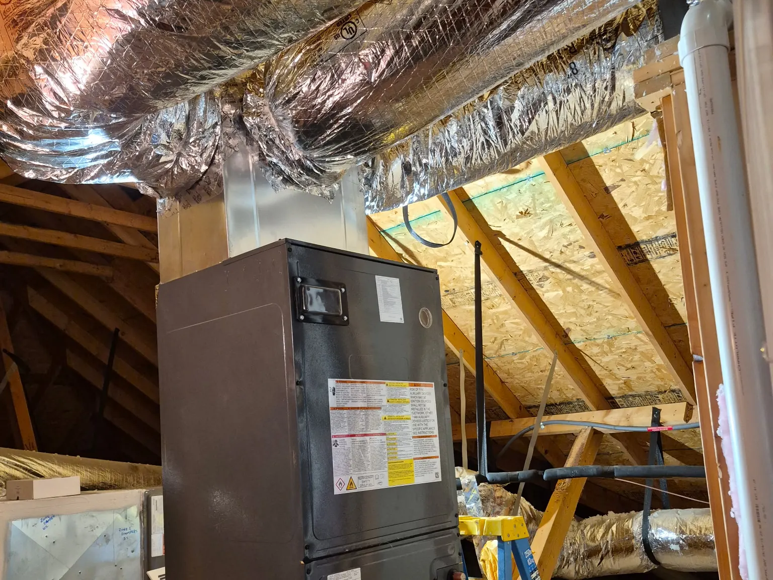 Upright air handler in an unfinished attic, under foil-wrapped supply ducts, with a step ladder beside it
