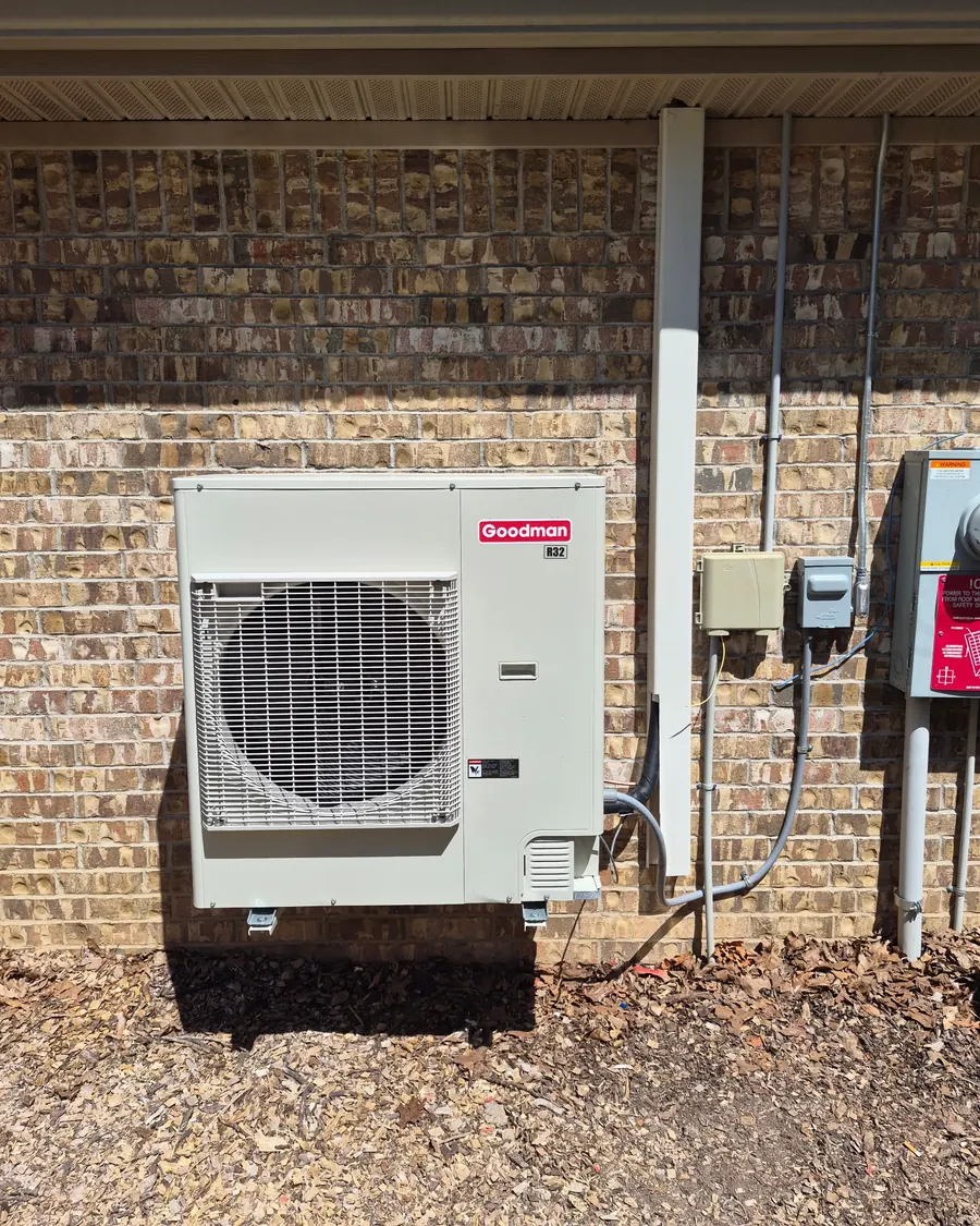 Goodman outdoor HVAC unit on wall brackets against a brick wall, with its disconnect box and electrical conduit beside it