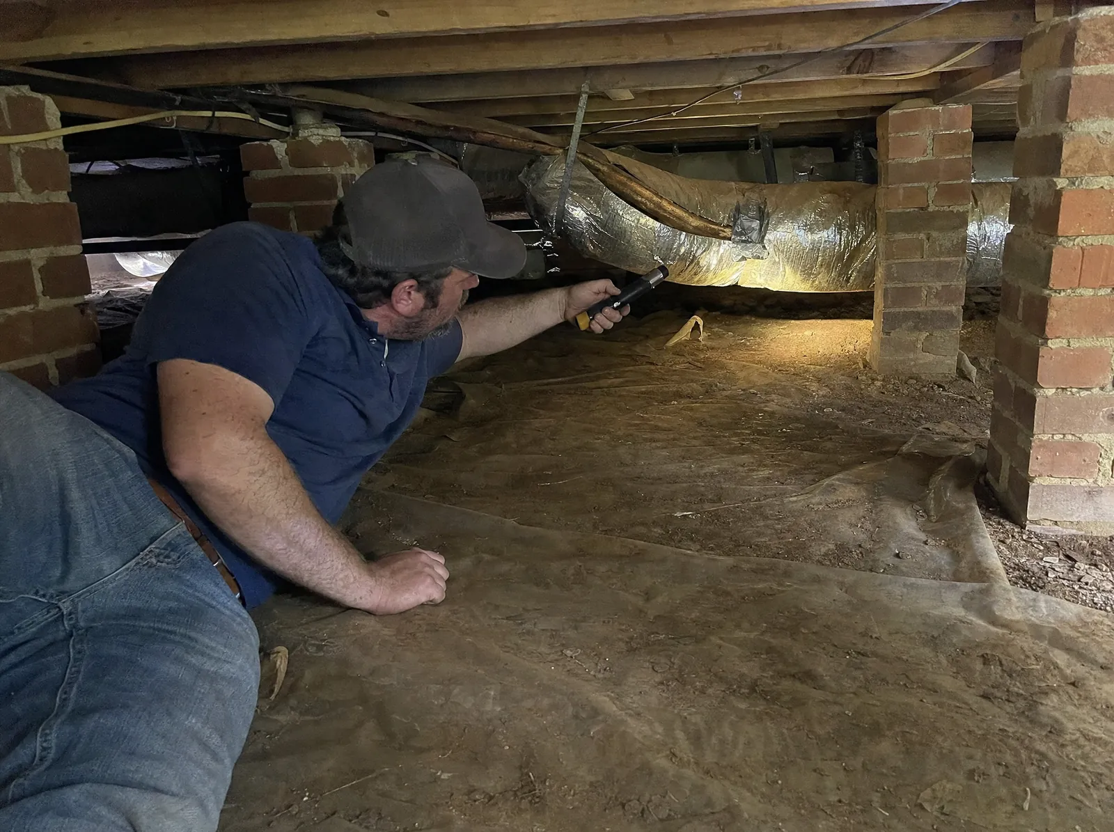 HVAC technician lying in a crawlspace, shining a flashlight along an insulated duct run between brick piers