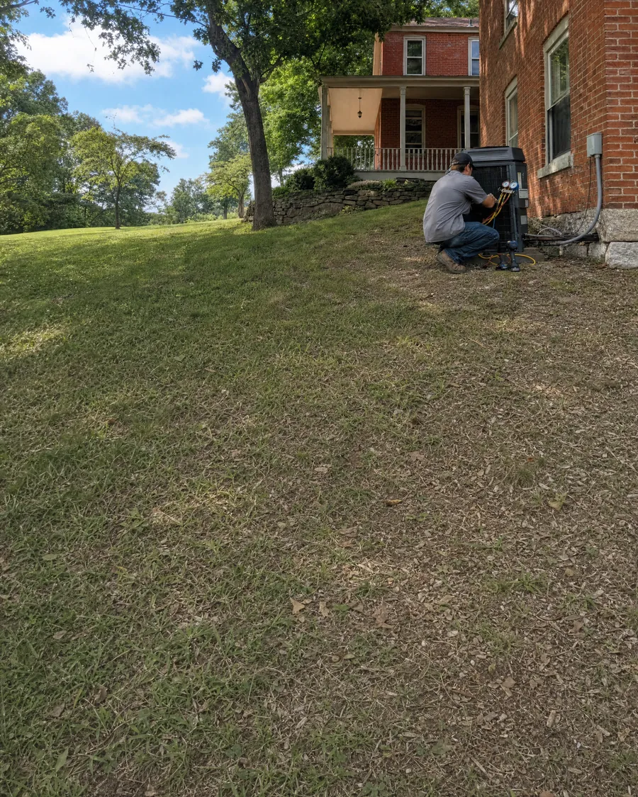 HVAC technician connecting refrigerant gauges to an outdoor air conditioner beside an older two-story red brick home with a wooden porch