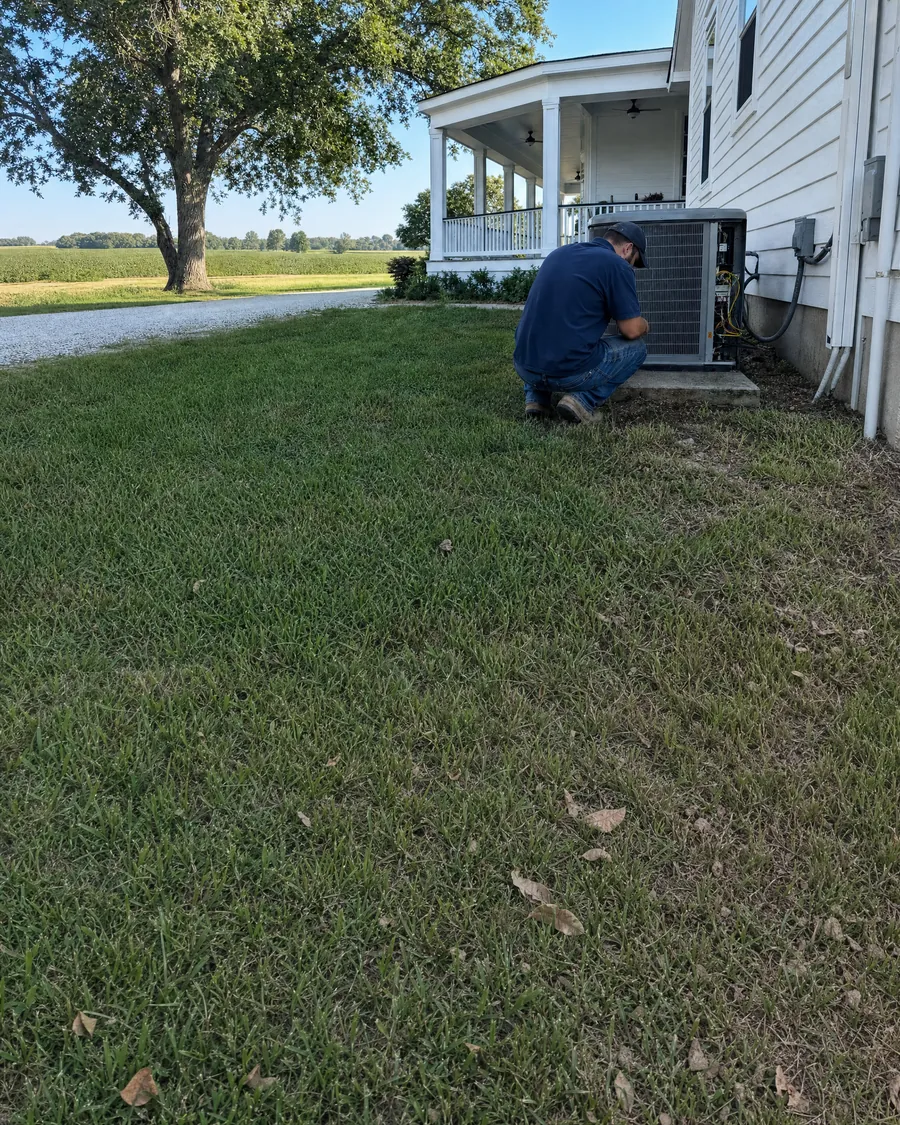 HVAC technician crouched at an outdoor air conditioner beside a white farmhouse with a wraparound porch and a large old oak tree
