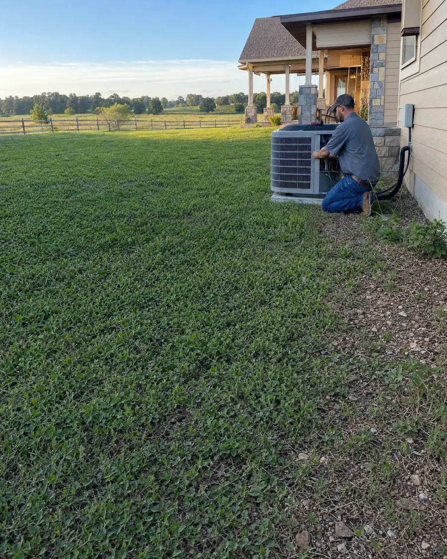 HVAC technician kneeling at an outdoor air conditioner beside a ranch home with stone accents, with a split rail fence and open fields beyond