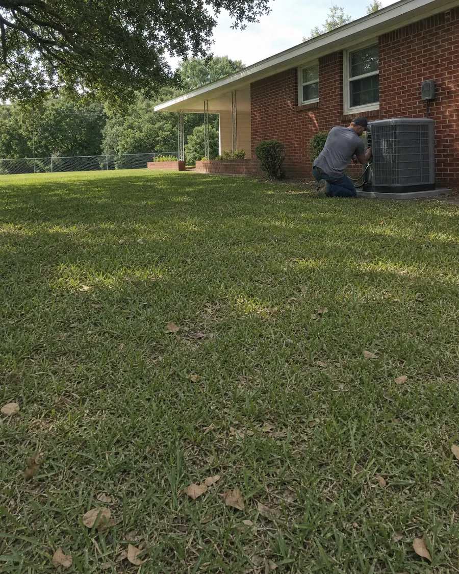 HVAC technician kneeling at an outdoor air conditioner beside a red brick ranch home with a carport and a large shade tree
