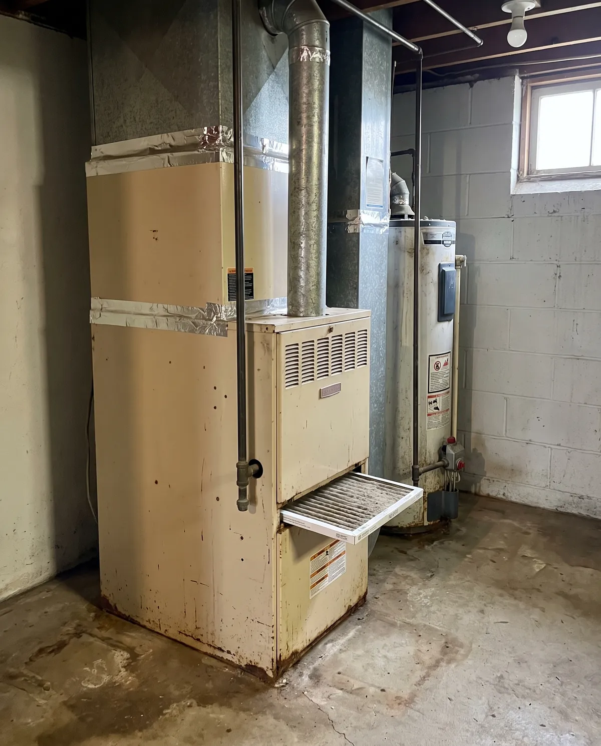 Older gas furnace with a rusted base and a dirty filter half pulled out, beside a water heater in an unfinished basement