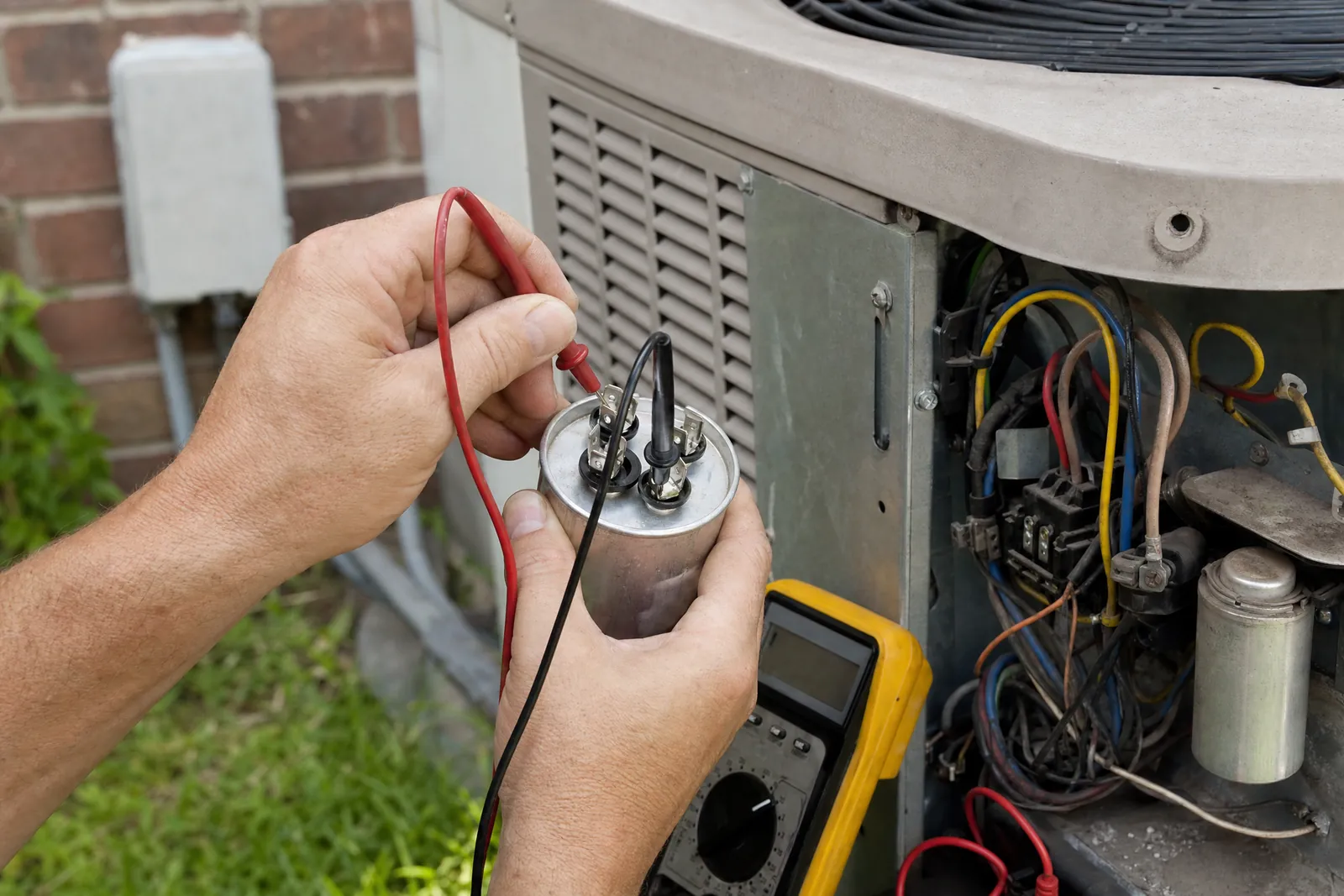 Two hands testing a run capacitor with a multimeter at an open condenser panel.